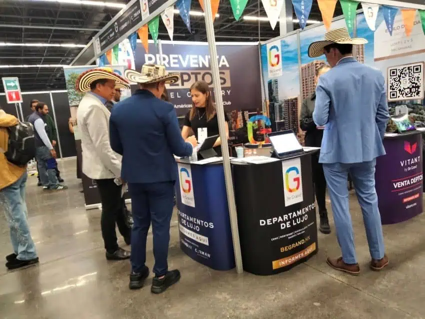 Three men wearing large, wide-brimmed, striped hats are visible from behind and face a booth counter in a large hall while women working the booth attend to them. The booth has two branded cylindrical stands and a backdrop banner with text including "PREVENTA DEPTOS" and "DEPARTAMENTO DE LUJO" and a large "G" logo. On the far right, another person in a light blue suit and a similar hat stands facing away from the camera, next to a display with a QR code.
