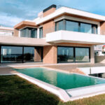 Want to buy property in Mexico? Check this guide to Mexico’s real estate expos first A bright, full exterior shot showcases a large, modern luxury home with a rectangular infinity pool in the foreground. The house features a striking contemporary design with white and earth-toned facades, expansive dark-framed windows and sliding glass doors, and multiple levels. A grassy lawn surrounds the pool, and in the distance, other residential buildings and clear skies with scattered clouds are visible.