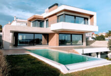 Want to buy property in Mexico? Check this guide to Mexico’s real estate expos first A bright, full exterior shot showcases a large, modern luxury home with a rectangular infinity pool in the foreground. The house features a striking contemporary design with white and earth-toned facades, expansive dark-framed windows and sliding glass doors, and multiple levels. A grassy lawn surrounds the pool, and in the distance, other residential buildings and clear skies with scattered clouds are visible.