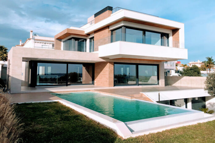 A bright, full exterior shot showcases a large, modern luxury home with a rectangular infinity pool in the foreground. The house features a striking contemporary design with white and earth-toned facades, expansive dark-framed windows and sliding glass doors, and multiple levels. A grassy lawn surrounds the pool, and in the distance, other residential buildings and clear skies with scattered clouds are visible.
