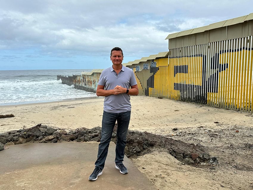 Travis Bembenek stands in front of the border wall in Tijuana where it meets the ocean