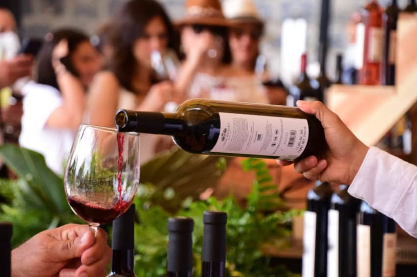 A close-up shot shows a person in a white long-sleeved shirt pouring red wine from a green bottle into a glass held by another person. In the soft-focus background, several people are visible, and more wine bottles are on display.