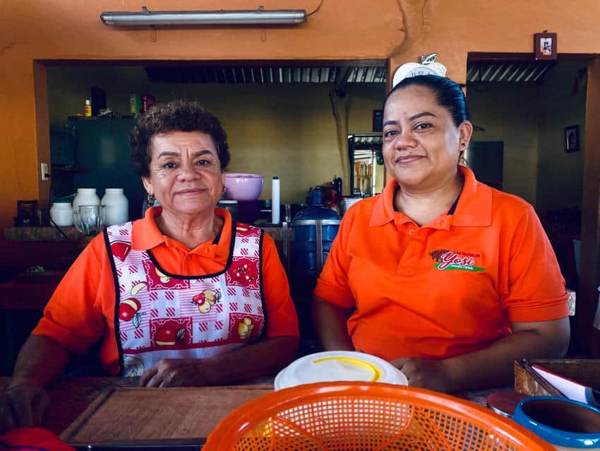 Two women, both wearing orange shirts, stand behind a counter in what appears to be a casual eatery or kitchen. The woman on the left wears a white apron with a colorful pattern and has short, curly hair. The woman on the right has her hair pulled back and a slight smile. Kitchen elements are visible in the background.