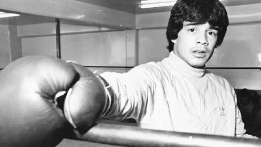 A black and white close-up shot of a young boxer in a training ring. He has dark, wavy hair and is wearing a light-colored, long-sleeved turtleneck or mock-neck shirt. His boxing gloved hand is prominently in the foreground, resting on a bar or rope, while his gaze is directed slightly off-camera with a focused expression. The background appears to be an indoor gym with a mirror and flourescent lighting fixtures.